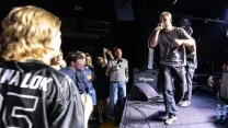 Wokalista z mikrofonem na scenie podczas występu na żywo, energiczna publiczność blisko estrady, przed sceną plastikowe butelki z wodą, w tle sprzęt muzyczny i głośnik z napisem „brighter”.