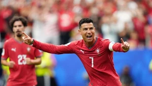 Piłkarz w czerwonym stroju z numerem 7 celebruje zdobycie gola, szeroko rozkładając ręce, na tle innych zawodników i kibiców na stadionie.