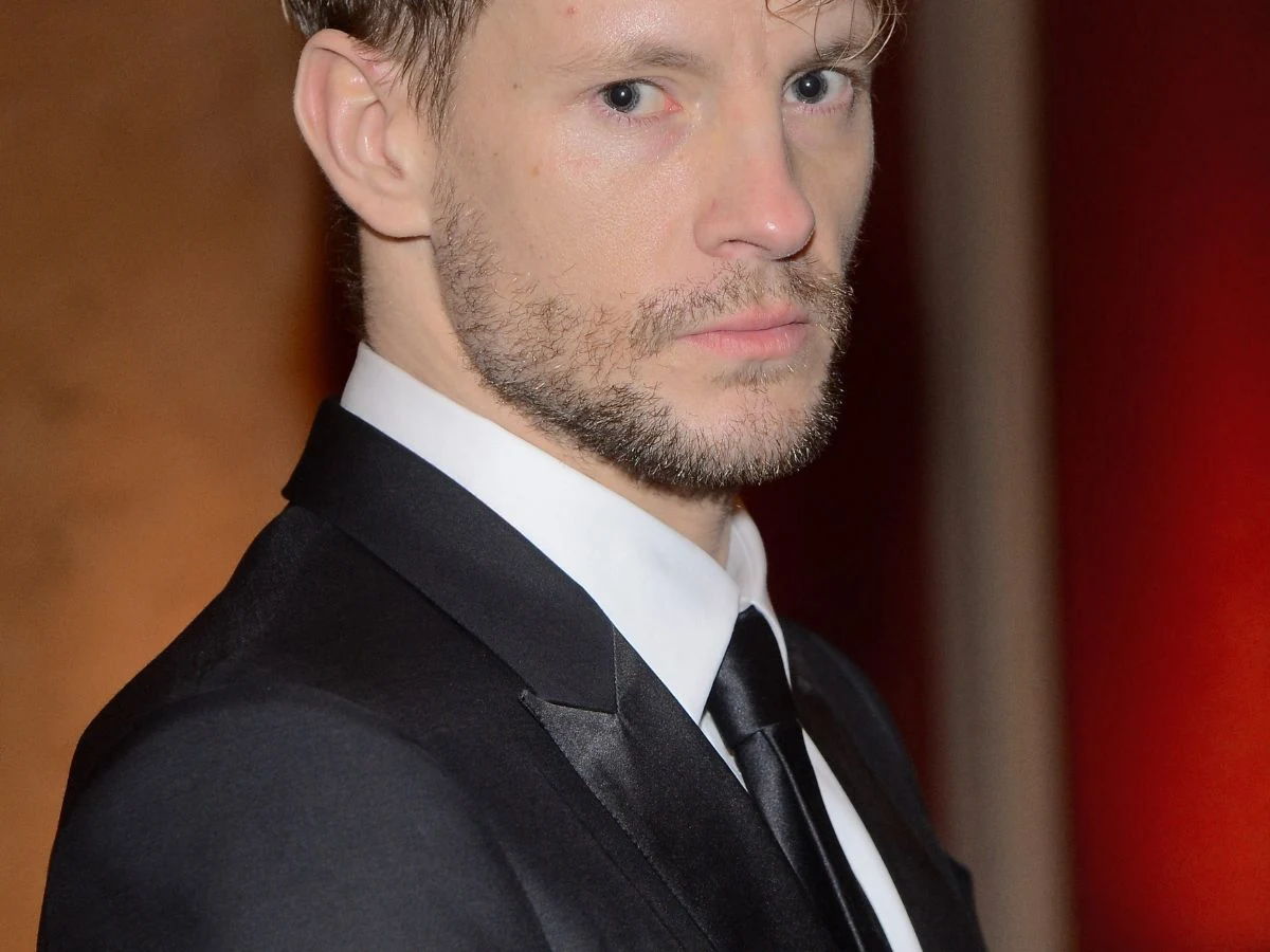 Sebastian Fabijański Sebastian Fabijański w eleganckim czarnym garniturze na czerwonym tle.