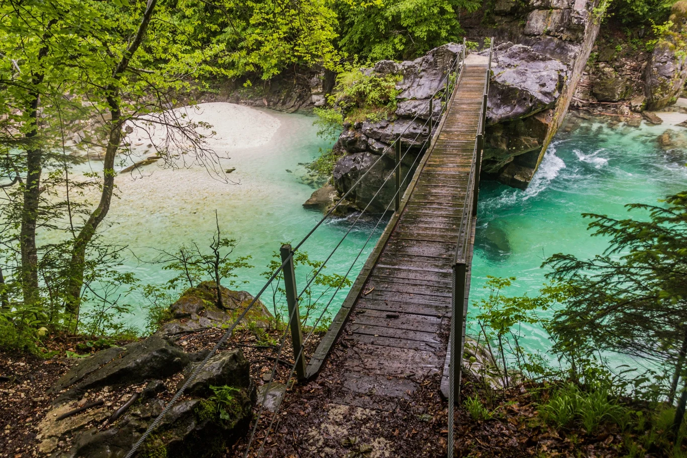 Drewniany most zawieszony nad turkusową rzeką otoczoną zielenią i skałami.