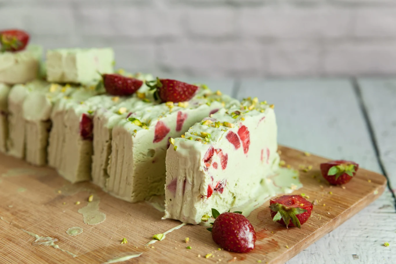 Pistachio ice cream with strawberry and pistachio nuts on a light wooden background Plastry semifreddo pistacjowego z truskawkami i posiekanymi pistacjami na drewnianej desce.