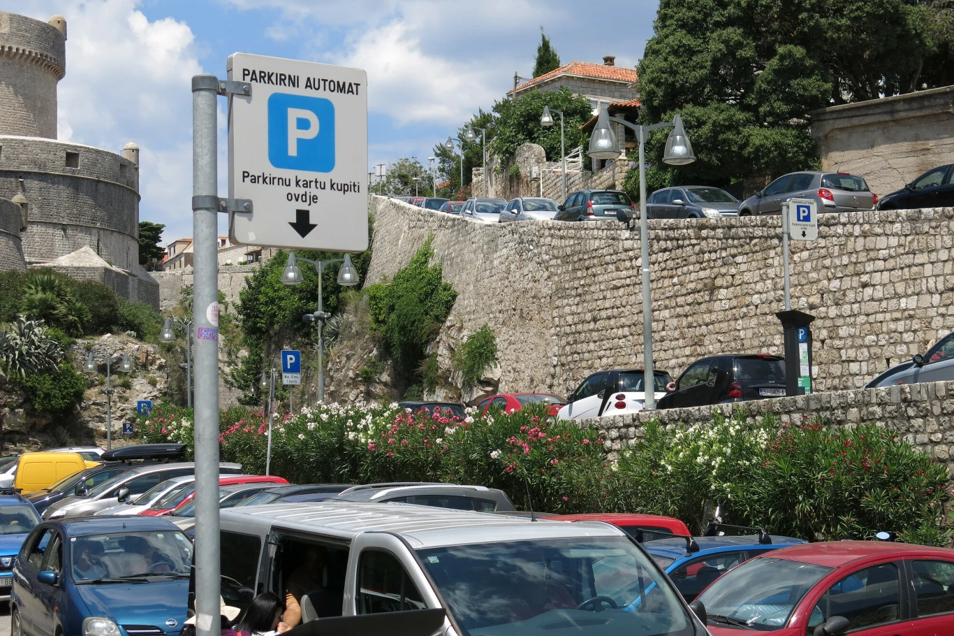 Znak parkingowy z napisem 'PARKIRNI AUTOMAT' oraz zaparkowane samochody na tle kamiennych murów i zieleni.