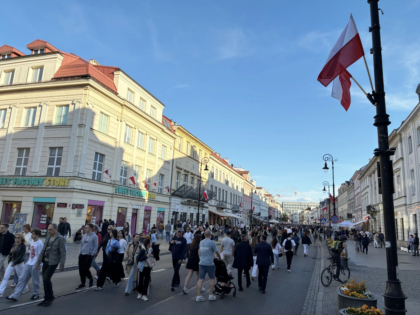 Tłum spacerujących ludzi na szerokiej ulicy otoczonej zabytkowymi kamienicami z biało-czerwonymi flagami powiewającymi na latarniach.