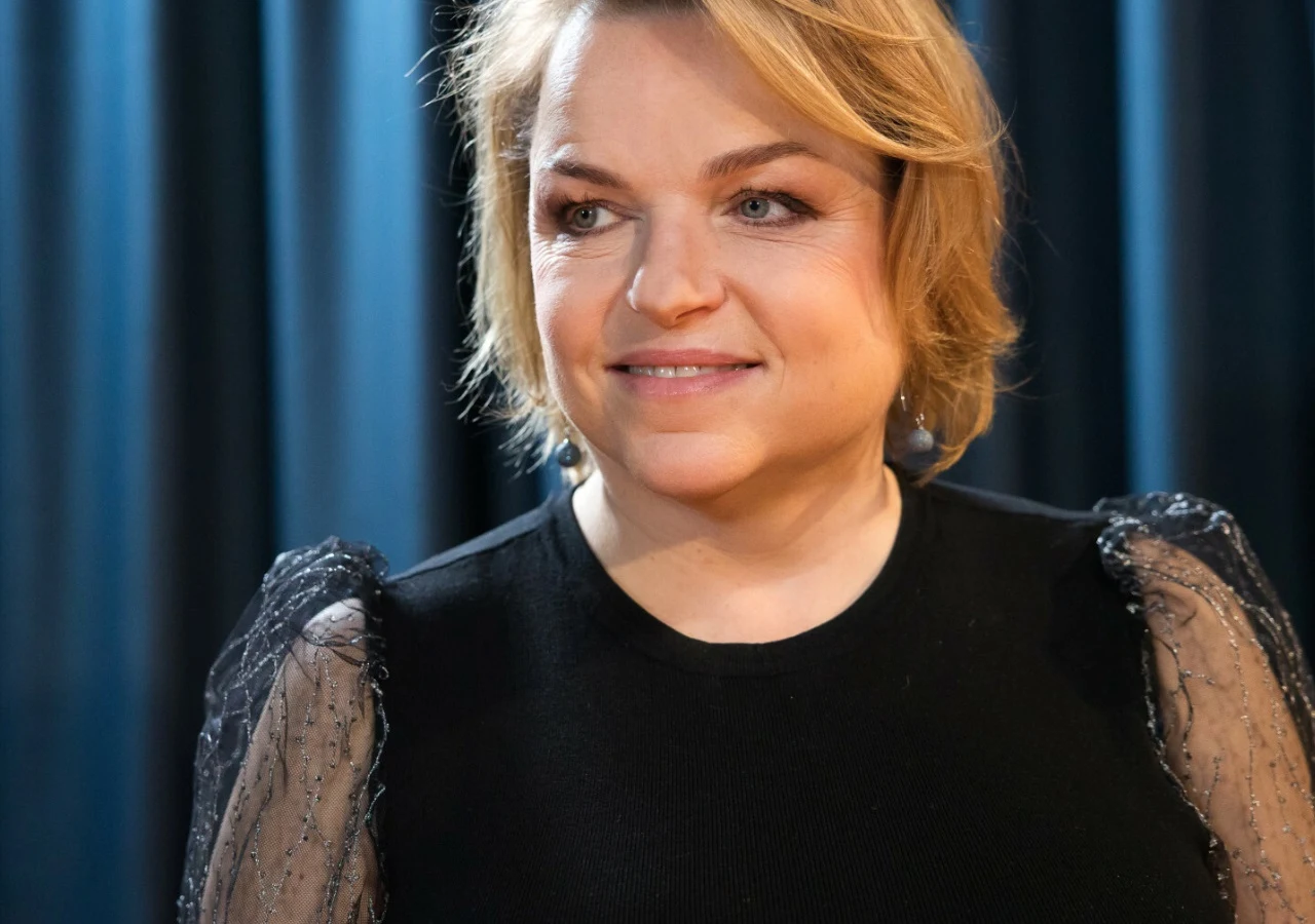 Katarzyna Bosacka Katarzyna Bosacka w czarnej bluzce z przeźroczystymi rękawami, stojąca na tle ciemnych zasłon.
