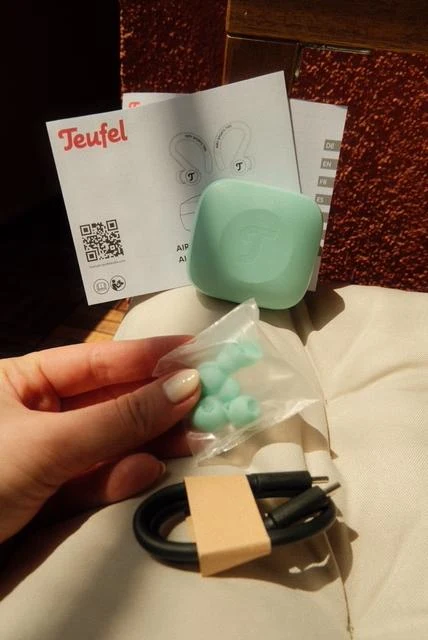 Zestaw słuchawek bezprzewodowych Teufel w pastelowozielonym etui, akcesoria w postaci wymiennych nakładek silikonowych oraz przewód do ładowania na tle instrukcji obsługi.