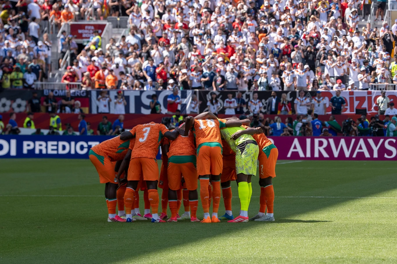 Grupa jedenastu piłkarzy w pomarańczowych strojach wraz z bramkarzem w jasnożółtym stroju tworzy krąg na murawie stadionu pełnego kibiców.