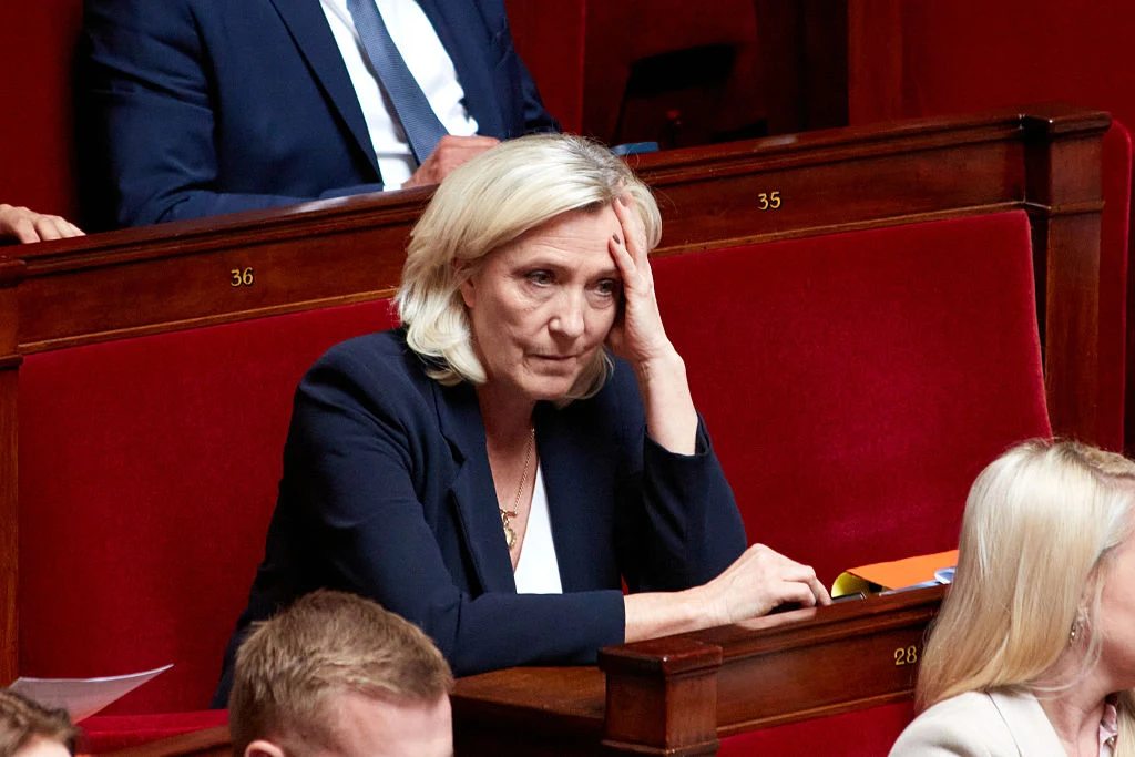 Liderka Zjednoczenia Narodowego Marine Le Pen Kobieta w średnim wieku z jasnymi włosami siedzi w czerwonym fotelu sali parlamentu, opierając głowę na dłoni i sprawiając wrażenie zamyślonej lub zmartwionej.