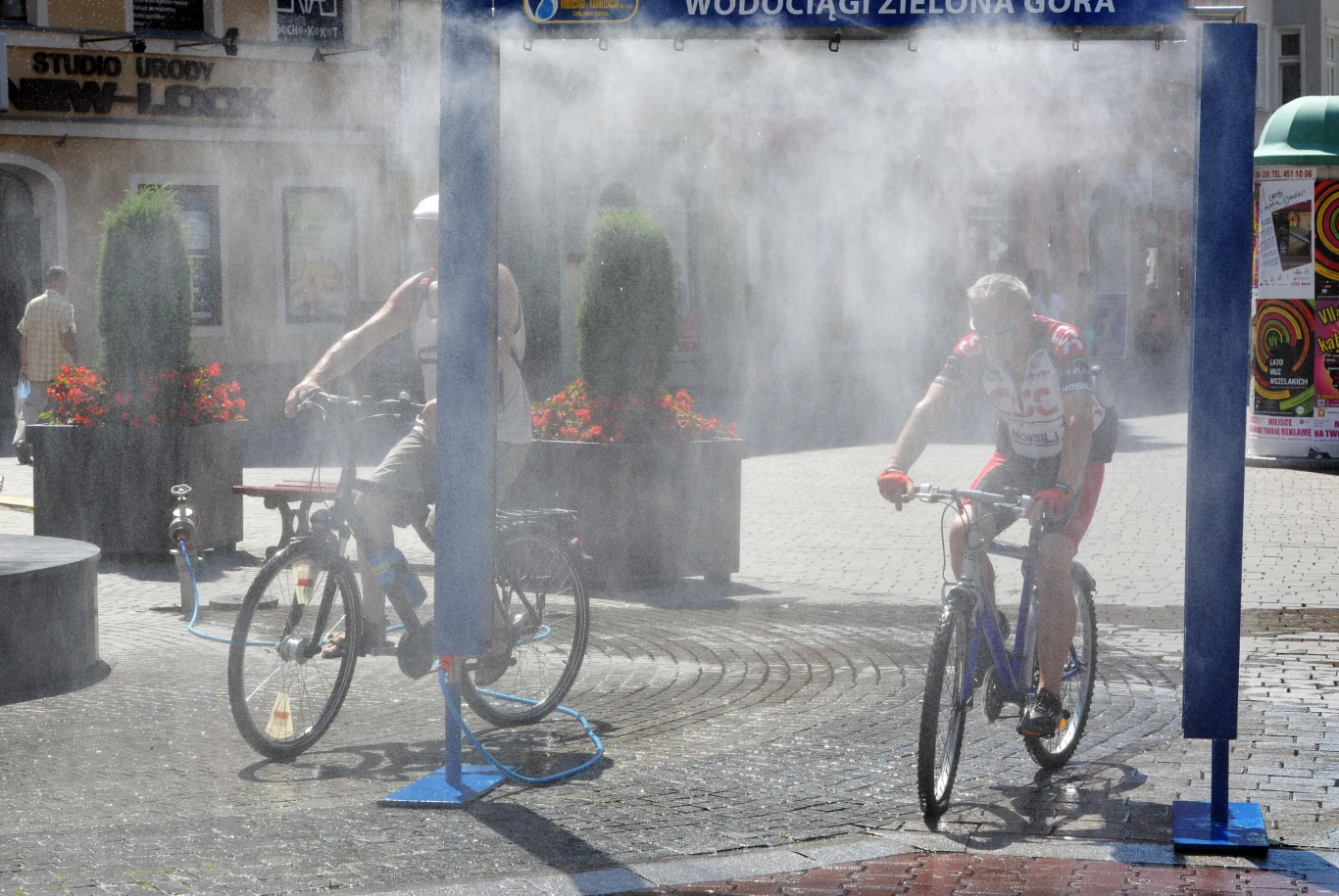 Dwóch rowerzystów przejeżdża przez kurtynę wodną na miejskim deptaku, korzystając z orzeźwienia podczas upalnego dnia.