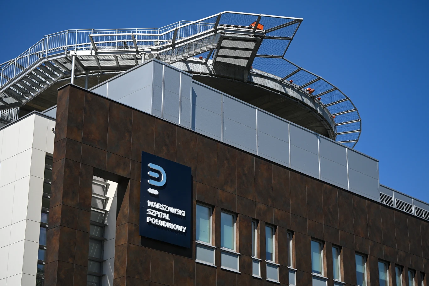 Nowoczesny budynek z elewacją w odcieniach brązu i bieli, logo oraz nazwa 'Warszawski Szpital Południowy' na jasnym tle, nad nimi platforma ewakuacyjna na dachu otoczona metalowymi barierkami.