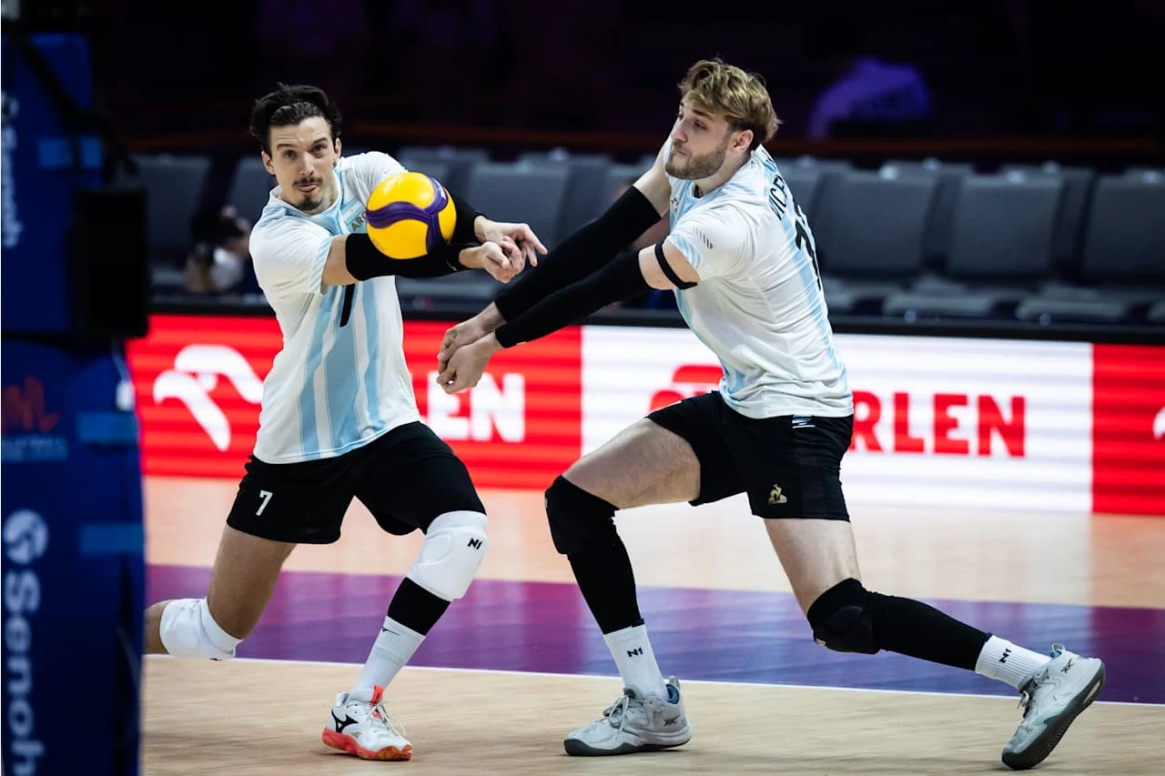 Dwóch siatkarzy w biało-niebieskich koszulkach skupionych na odbiorze piłki podczas meczu na hali sportowej.