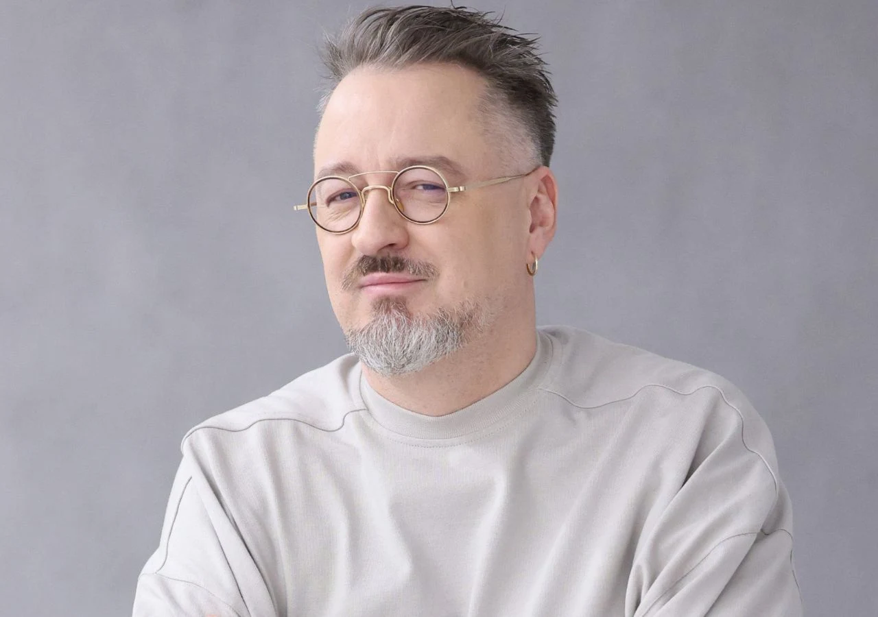 Piotr Kędzierski Piotr Kędzierski z zarostem i w okularach, ubrany w białą bluzę, siedzi na jasnym tle