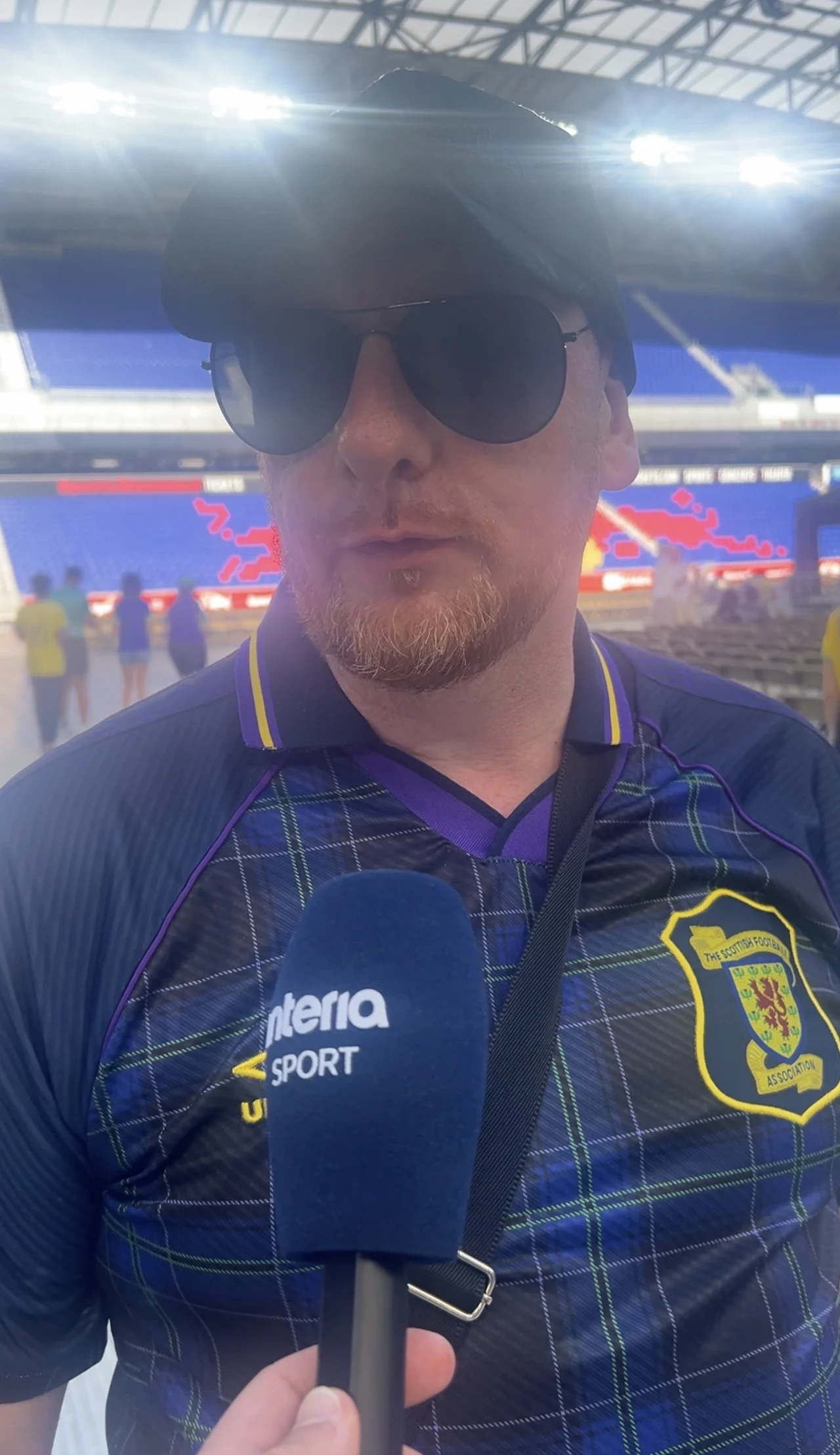 Mężczyzna w ciemnych okularach i czapce z daszkiem udziela wywiadu przed mikrofonem Interia Sport, ubrany w koszulkę z emblematem i wzorem przypominającym barwy szkockiej drużyny piłkarskiej, na tle stadionu piłkarskiego.