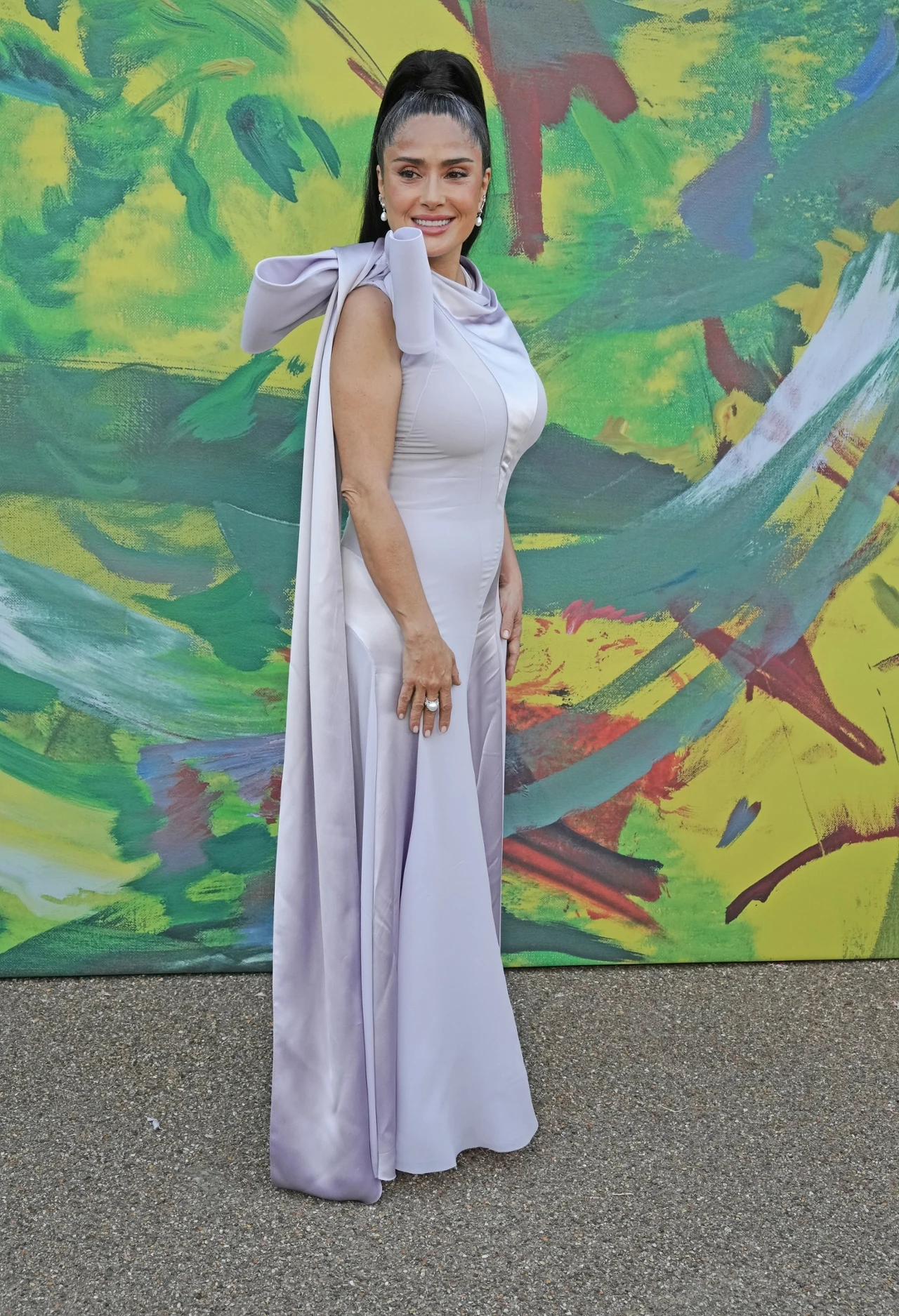 Salma Hayek wybrała niezwykle elegancką i zmysłową kreację na tę wyjątkową okazję Kobieta w długiej, jasnofioletowej sukni z efektowną kokardą na ramieniu pozuje na tle kolorowego, abstrakcyjnego muralu.