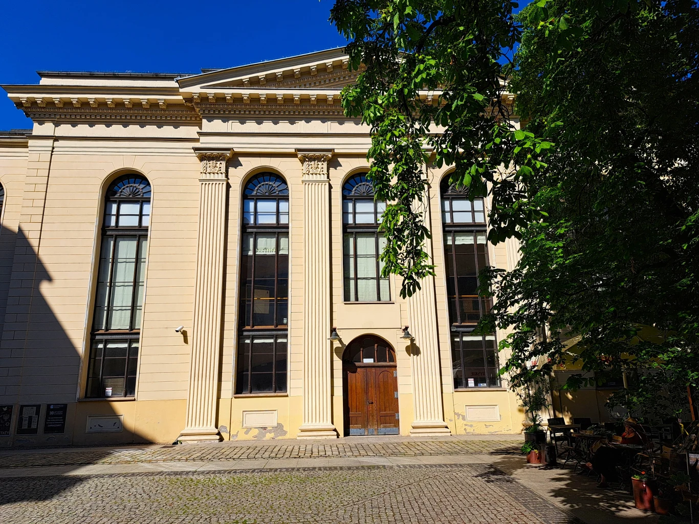 Synagoga pod Białym Bocianem skryta jest w podwórku Klasycystyczna fasada budynku z trzema wysokimi oknami zakończonymi łukiem, ozdobnymi kolumnami i masywnymi drzwiami drewnianymi pośrodku; na pierwszym planie brukowany plac, po prawej fragment ogródka kawiarnianego w cieniu drzew.
