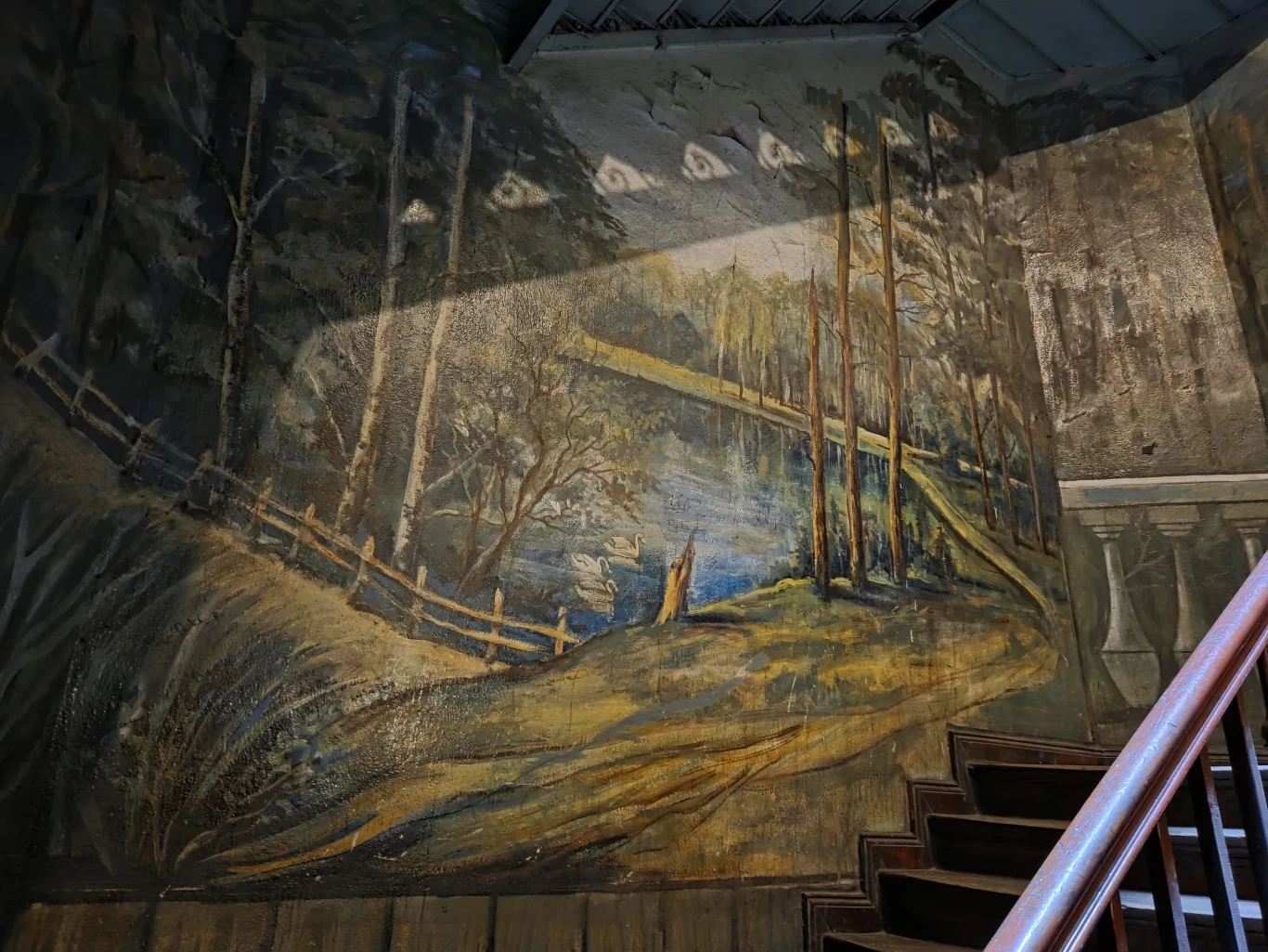 Klatka schodowa na ulicy Glinianej 60 we Wrocławiu Malowidło ścienne przedstawia drewnianą kładkę przerzuconą nad rzeką w lesie, otoczoną wysokimi drzewami oraz drewnianym płotem, z efektownym wykorzystaniem światłocienia.