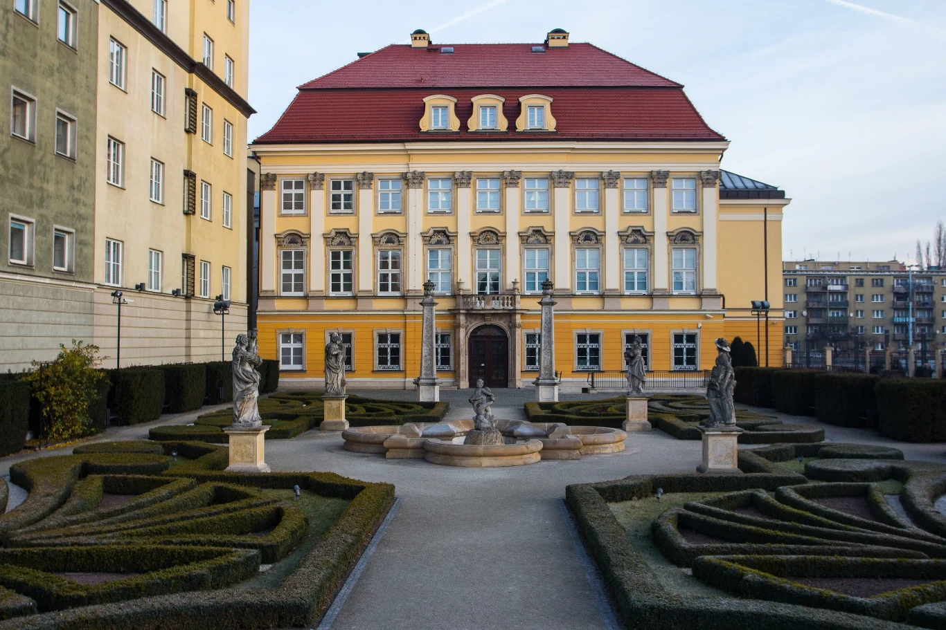 Pałac Królewski we Wrocławiu dziś jest siedzibą miejskiego muzeum Barokowy pałac z żółtą fasadą otoczony symetrycznym ogrodem francuskim, w którym stoją kamienne rzeźby i fontanna pośrodku alejek z geometrycznie przyciętymi krzewami.