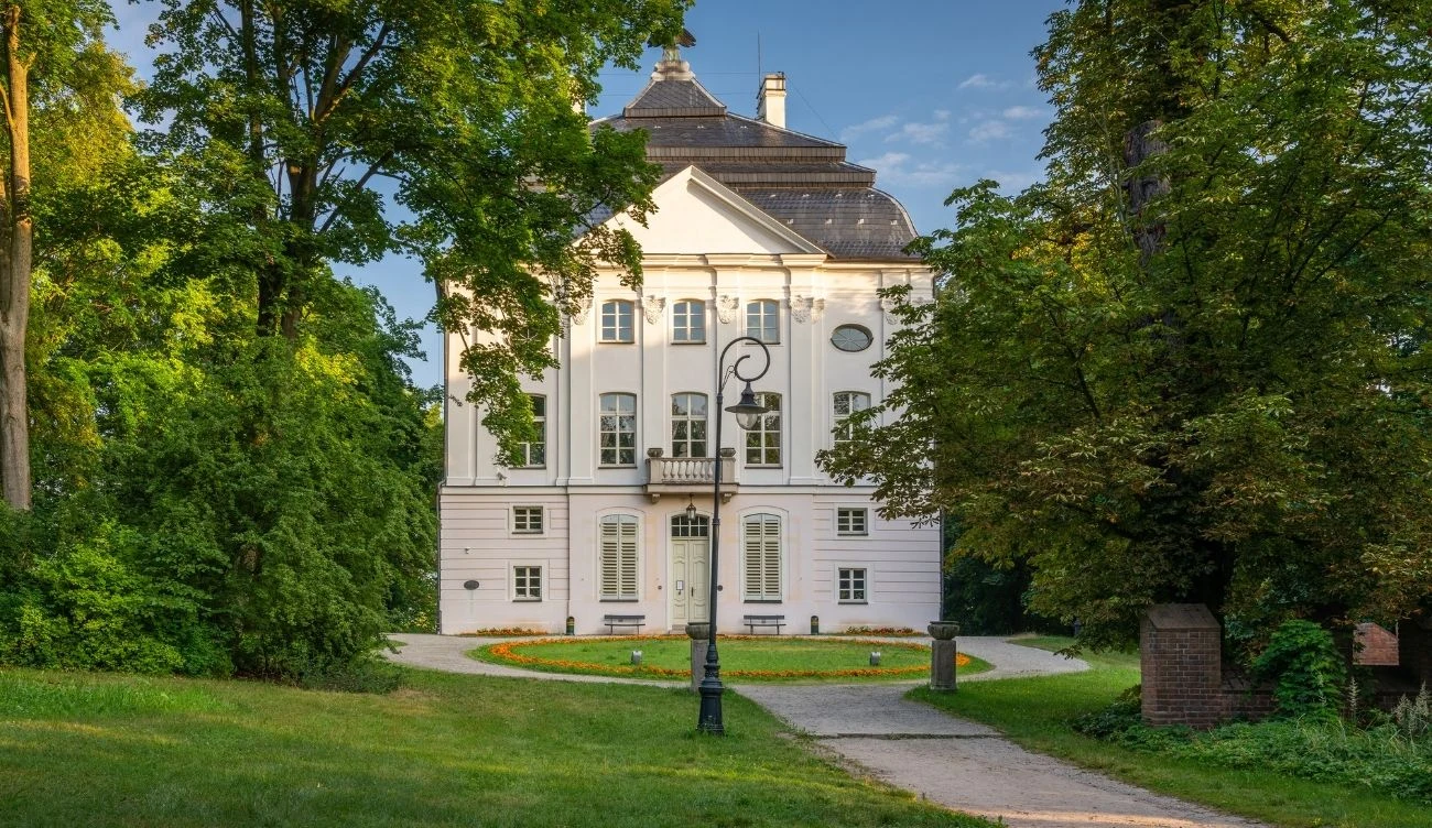 Biała, klasycystyczna rezydencja otoczona bujną zielenią, stojąca przy ścieżce prowadzącej przez park, ozdobiona balkonem, kolumnami oraz wysokimi oknami, z centralnie ustawioną stylową latarnią.