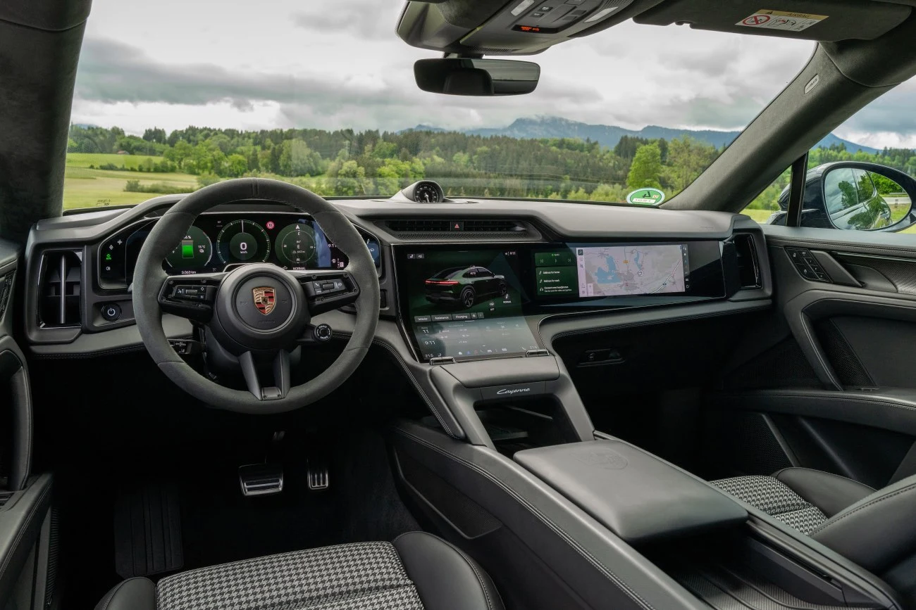 Luksusowe wnętrze samochodu Porsche z nowoczesnym kokpitem, wielofunkcyjną kierownicą, cyfrowymi wyświetlaczami oraz rozbudowanym panelem centralnym, a za oknami rozpościera się widok na zielone pola i góry.