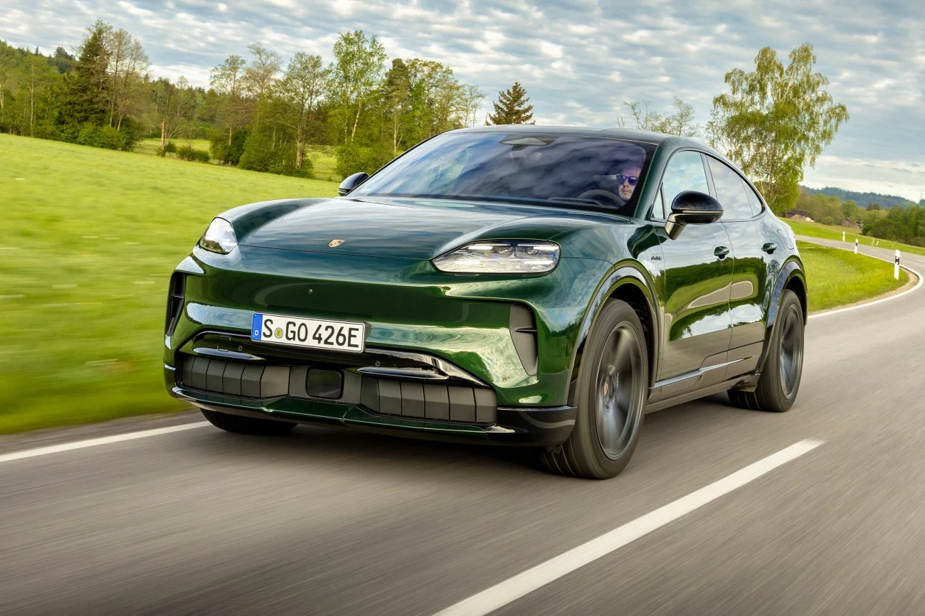 Zielony samochód marki Porsche w dynamicznym ruchu na krętej, asfaltowej drodze, otoczonej zielonymi polami i drzewami.