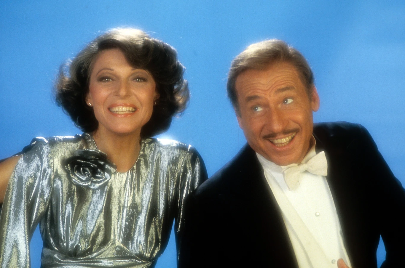 Anne Bancroft i Mel Brooks w filmie "Być albo nie być" (1983) / 20th Century-Fox Uśmiechnięta para w eleganckich, błyszczących strojach na jednolitym, niebieskim tle; kobieta w srebrnej sukni z kwiatową broszką na ramieniu, mężczyzna w czarnym smokingu z białą muchą.
