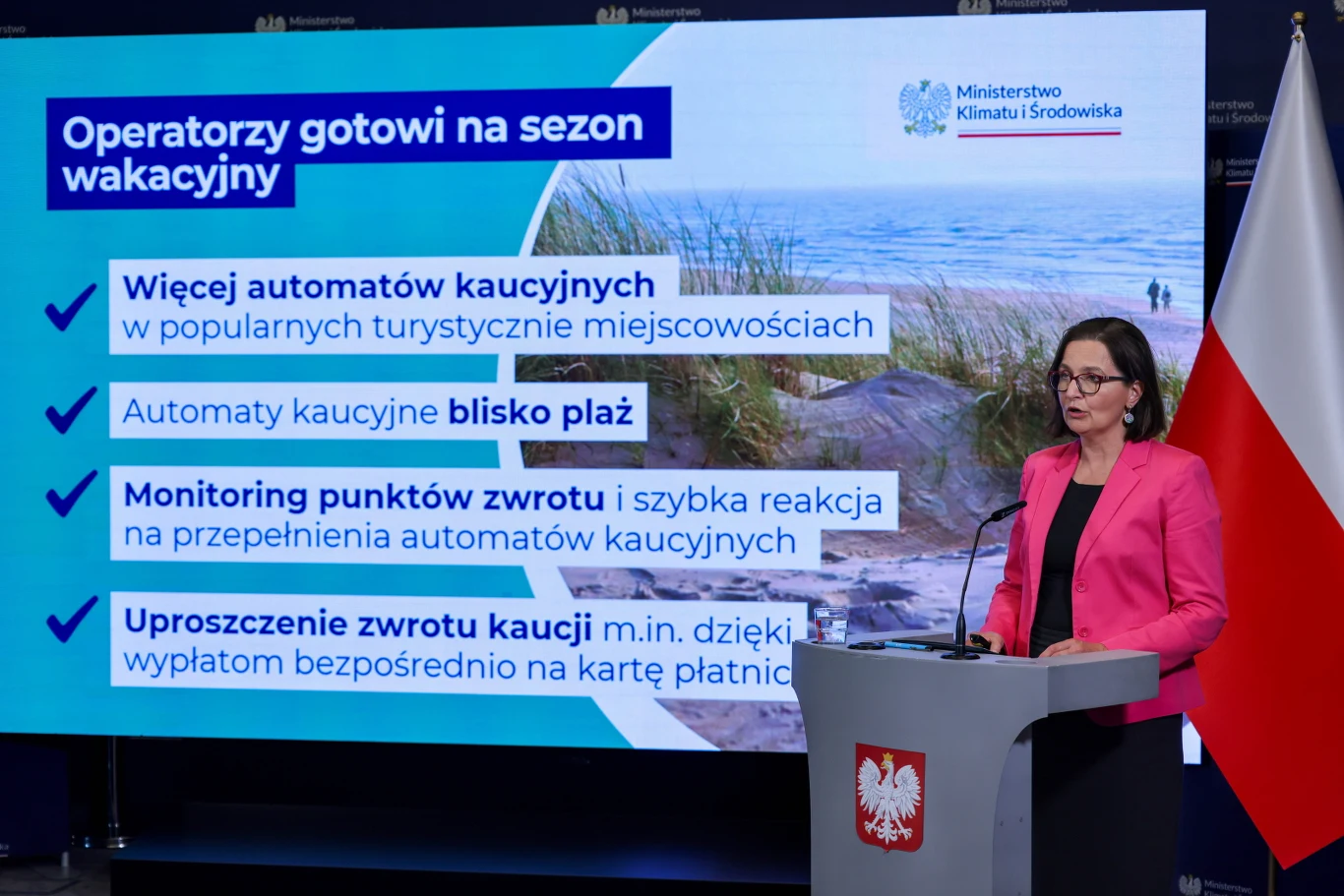 Kobieta w różowej marynarce wygłasza prezentację na tle dużego ekranu, na którym widoczne są kluczowe informacje dotyczące wdrożenia automatów kaucji oraz usprawnienia systemu zwrotu kaucji w popularnych turystycznych miejscowościach, a obok niej znajd...