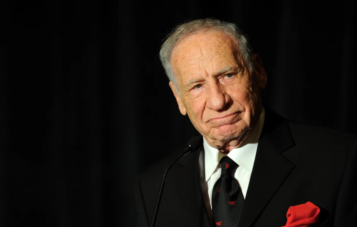 Mel Brooks ma już 100 lat Mel Brooks w czarnym garniturze, białej koszuli i ciemnym krawacie, stoi przy mikrofonie.