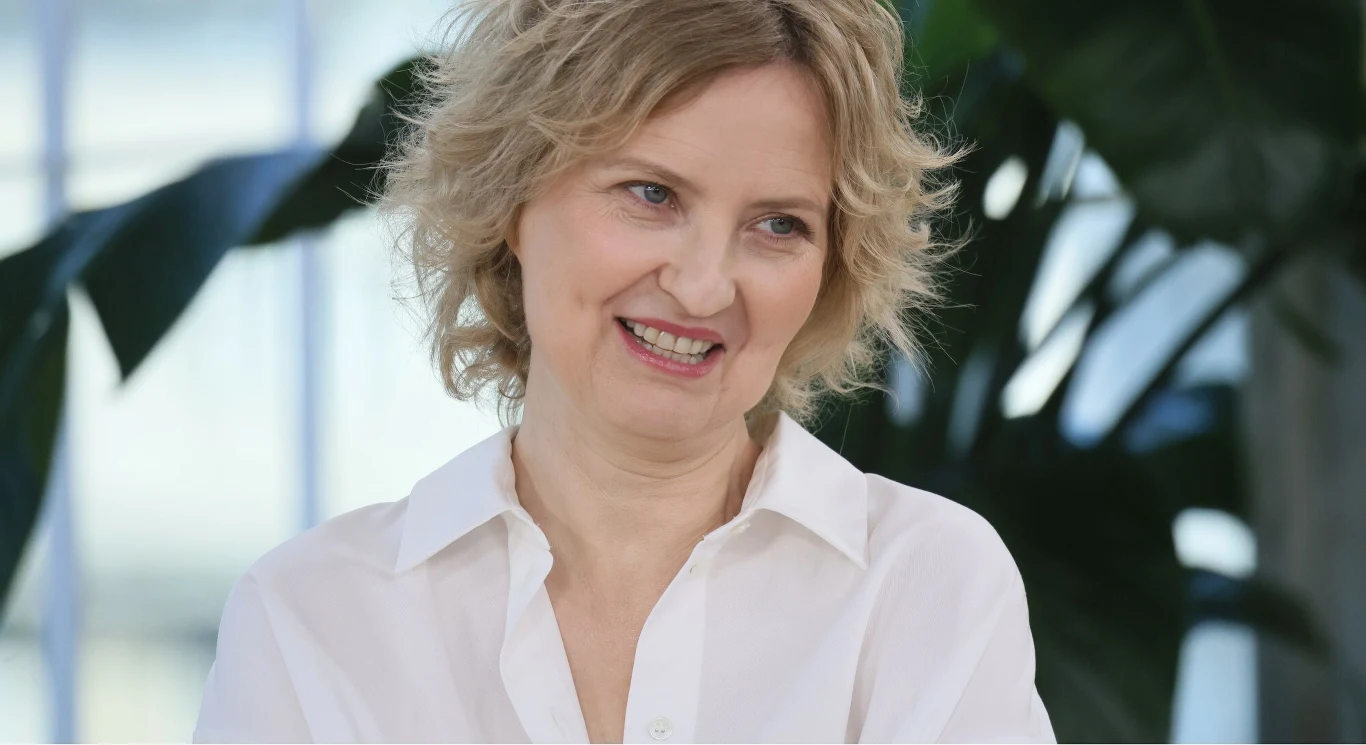 Izabela Kuna Izabela Kuna w białej koszuli, jasne kręcone włosy, w tle rozmyte zielone liście roślin.