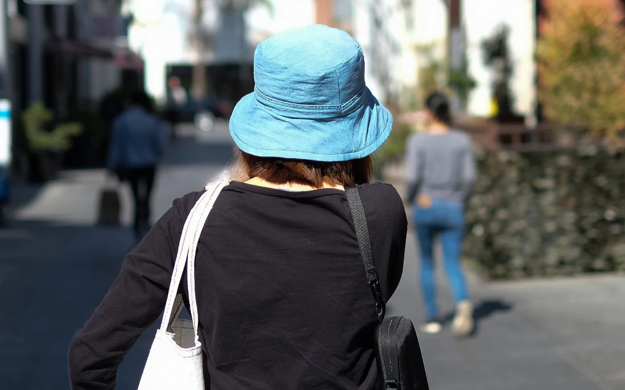 Osoba w niebieskim kapeluszu i czarnej bluzie idzie chodnikiem w słoneczny dzień, z torbą na ramieniu, w otoczeniu przechodzących ludzi.