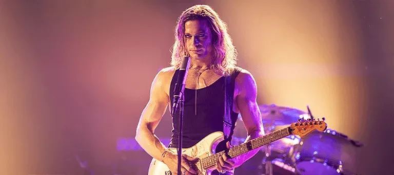 Mężczyzna z długimi blond włosami gra na gitarze elektrycznej i śpiewa do mikrofonu na scenie oświetlonej ciepłym światłem.