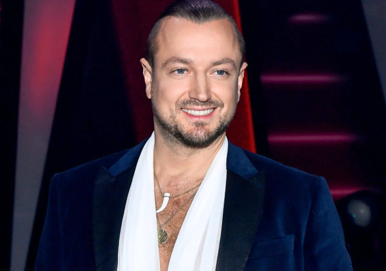 Aleksander Milwiw-Baron Uśmiechnięty mężczyzna w granatowej marynarce i białym szaliku, z krótkimi, zaczesanymi do tyłu włosami oraz złotym łańcuszkiem na szyi.