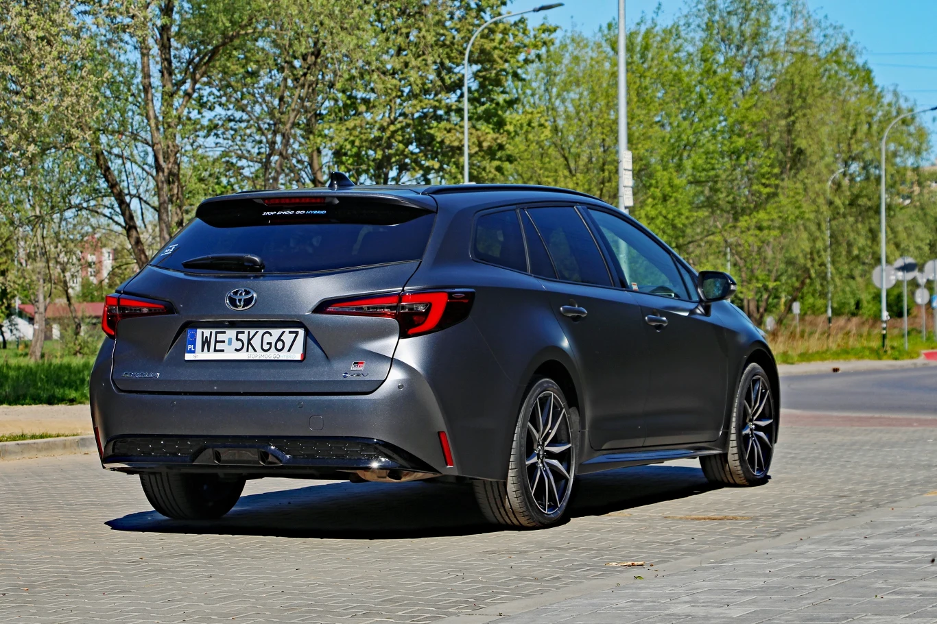 Ciemnoszara Toyota Corolla kombi z nowoczesnymi felgami, widziana z tyłu, zaparkowana na wybrukowanym placu, otoczona zielenią drzew.