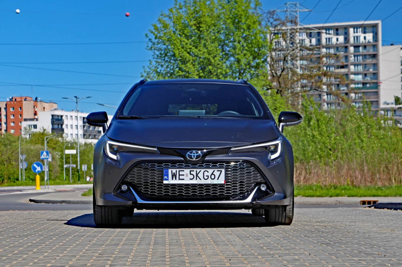 Czarny samochód Toyota z polską rejestracją, zaparkowany na kostce brukowej na tle zieleni i nowoczesnych budynków mieszkalnych.