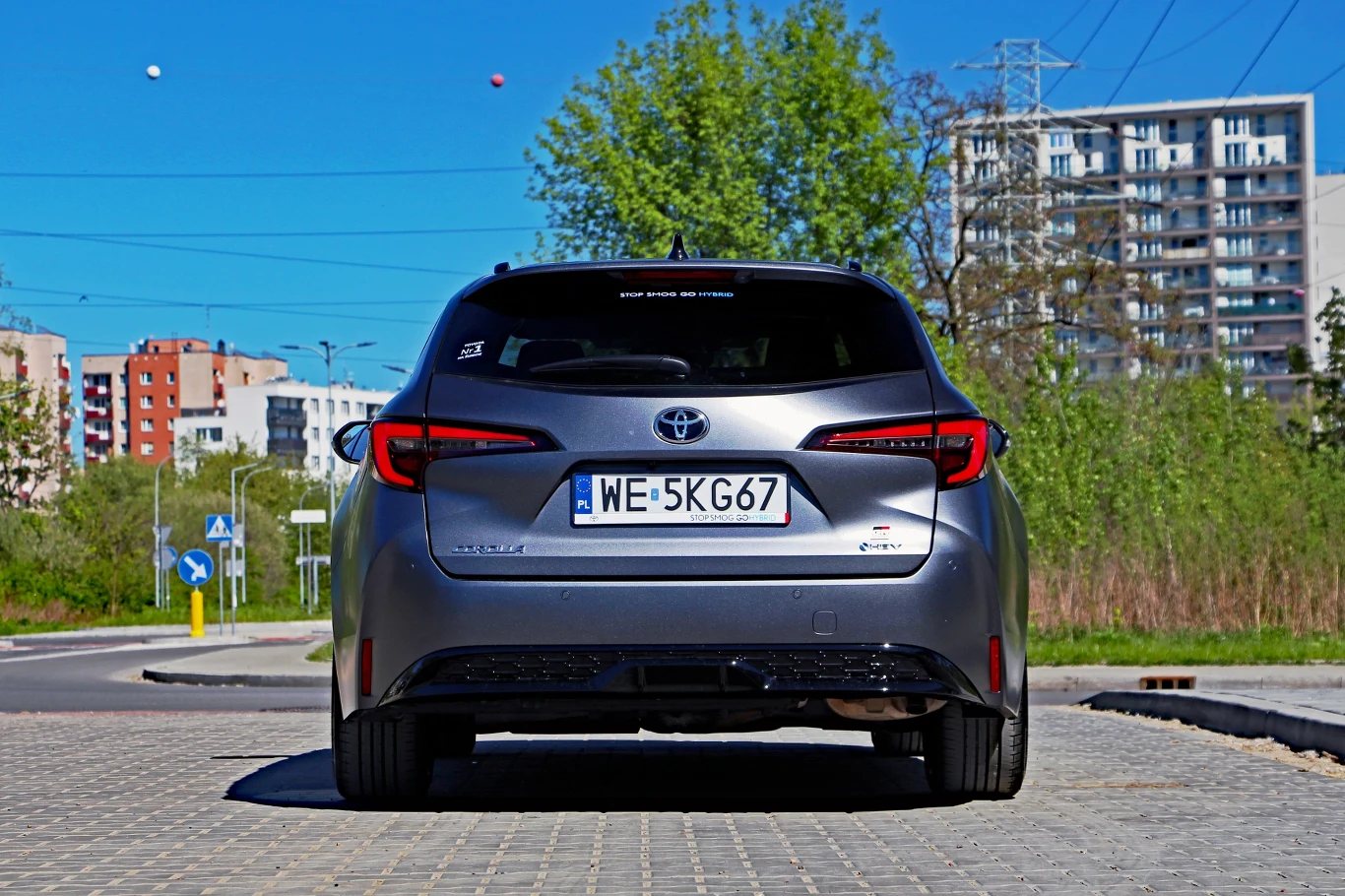 Srebrna Toyota z widoczną tablicą rejestracyjną, zaparkowana na tle zieleni oraz nowoczesnych bloków mieszkalnych.
