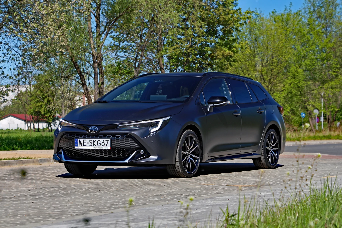 Czarna Toyota Corolla kombi zaparkowana na kostce brukowej, otoczona zielenią i drzewami w słoneczny dzień.