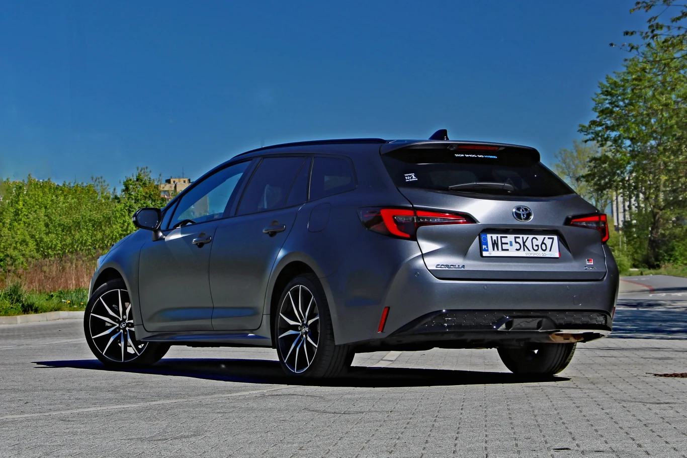 Szary samochód typu kombi marki Toyota zaparkowany na otwartym, wybrukowanym placu, otoczony zielenią i stojący na tle bezchmurnego nieba.