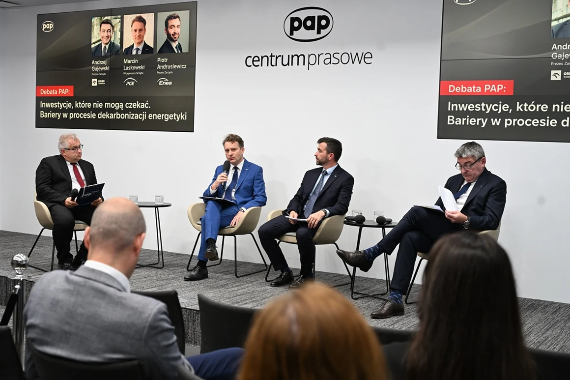 Czterech mężczyzn w garniturach siedzi na fotelach podczas panelu dyskusyjnego w centrum prasowym PAP, rozmawiając o barierach w procesie dekarbonizacji energetyki, a przed nimi znajduje się publiczność.