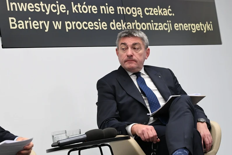 Mężczyzna w garniturze siedzi na krześle z teczką na kolanach, obok mikrofony i stolik, w tle baner z napisem dotyczącym barier w procesie dekarbonizacji energetyki.