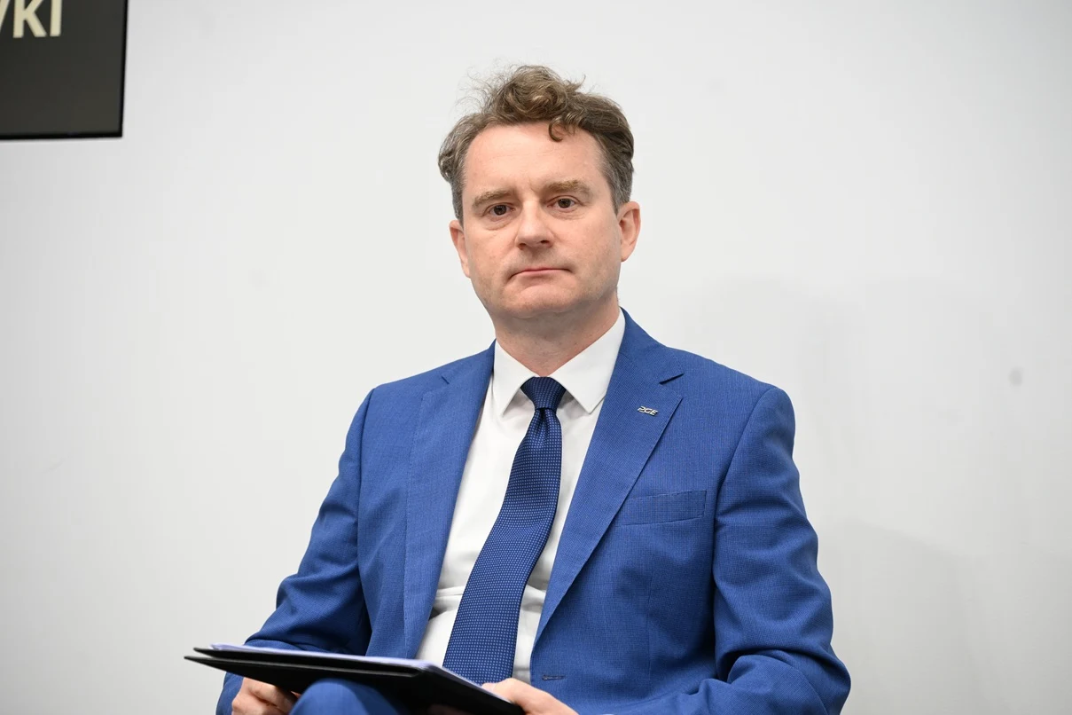 Mężczyzna w niebieskim garniturze i białej koszuli trzymający czarną teczkę, siedzący na tle jasnej ściany.