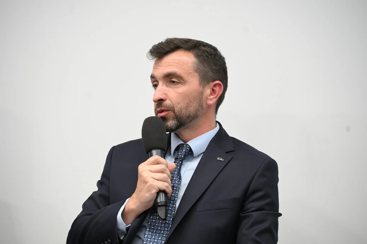 Mężczyzna w garniturze z krawatem, trzymający mikrofon i przemawiający, w tle jednolite jasne tło.