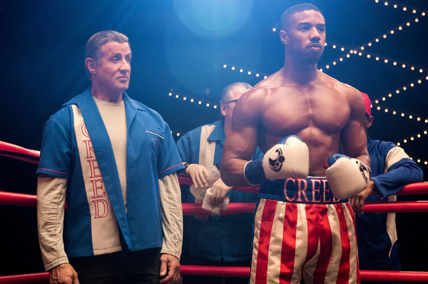 Umięśniony bokser w kolorowych spodenkach i rękawicach stoi w ringu obok starszego mężczyzny w sportowym ubraniu z napisem „Creed”.