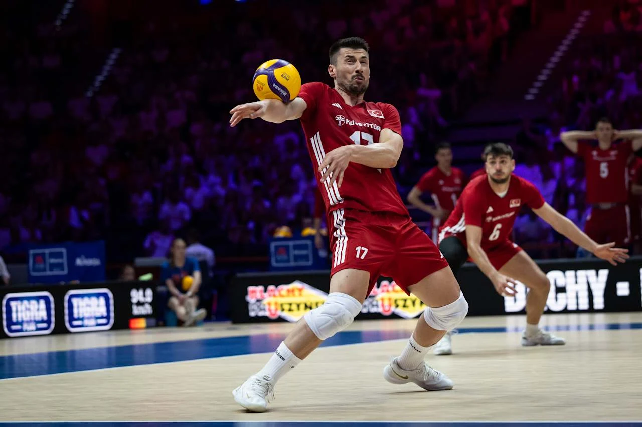 Murat Yenipazar Siatkarz w czerwonym stroju wykonujący przyjęcie piłki odbitej sposobem dolnym podczas meczu, w tle pozostali zawodnicy drużyny przygotowujący się do gry.