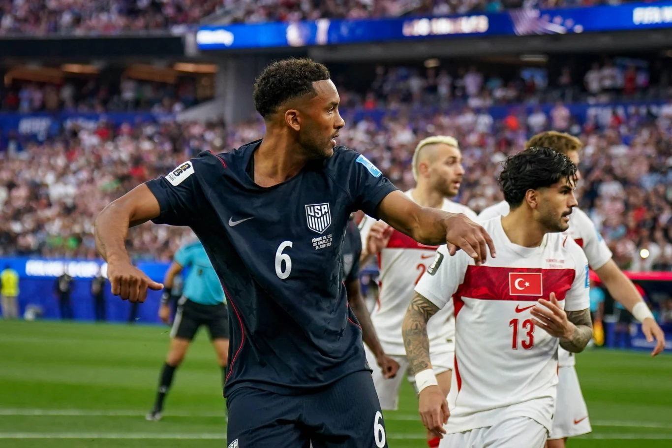 Piłkarz reprezentacji USA w ciemnogranatowym stroju z numerem 6 wykonuje dynamiczny ruch rękami podczas meczu, obok niego zawodnicy drużyny Turcji w białych koszulkach z czerwonym pasem obserwują przebieg akcji na boisku.