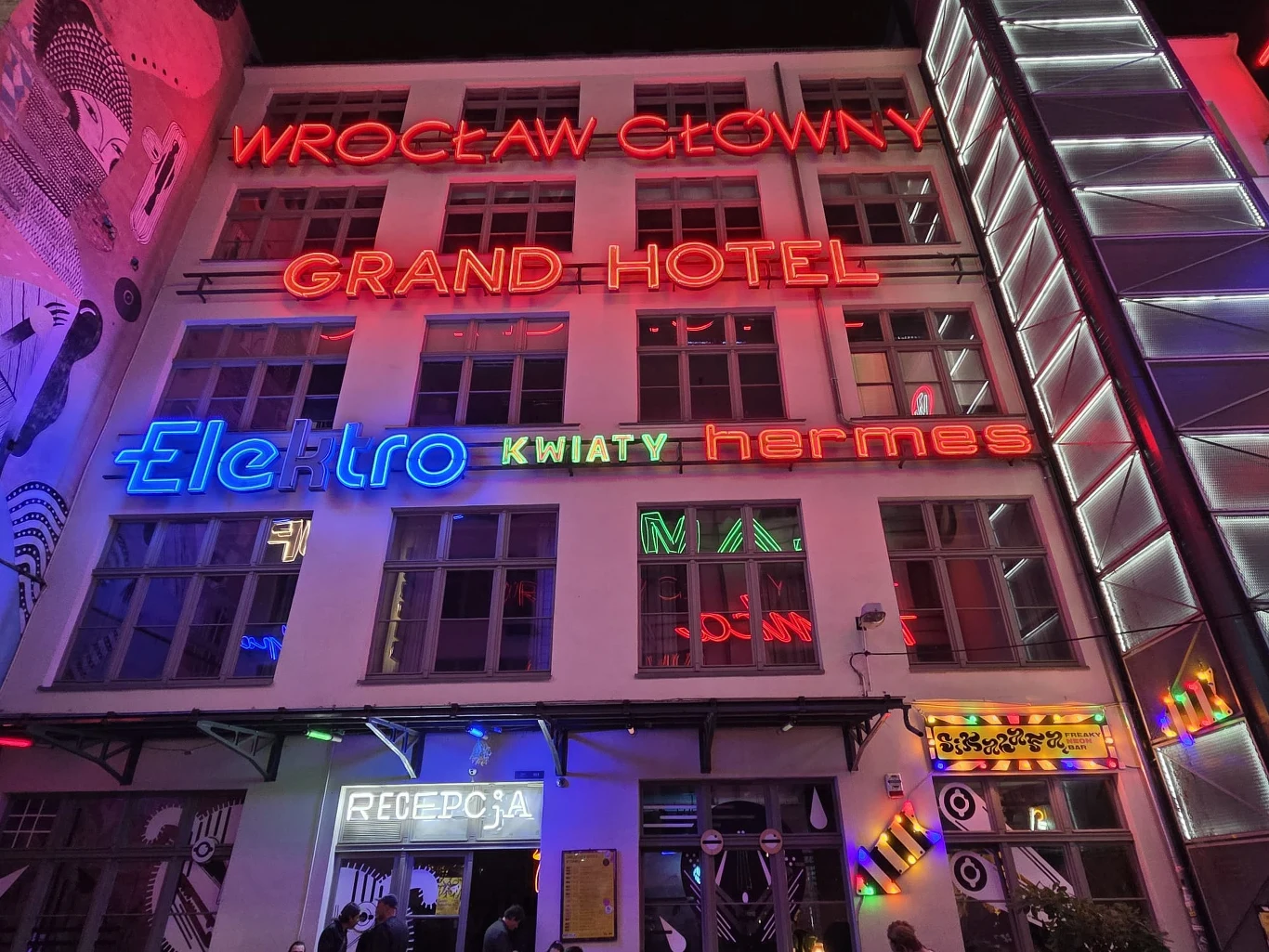 Fasada budynku ozdobiona kolorowymi neonowymi napisami, takimi jak Wrocław Główny, Grand Hotel, Elektro, Kwiaty, hermes; na dole wejście z szyldem recepcja oraz liczne światła w różnych barwach.