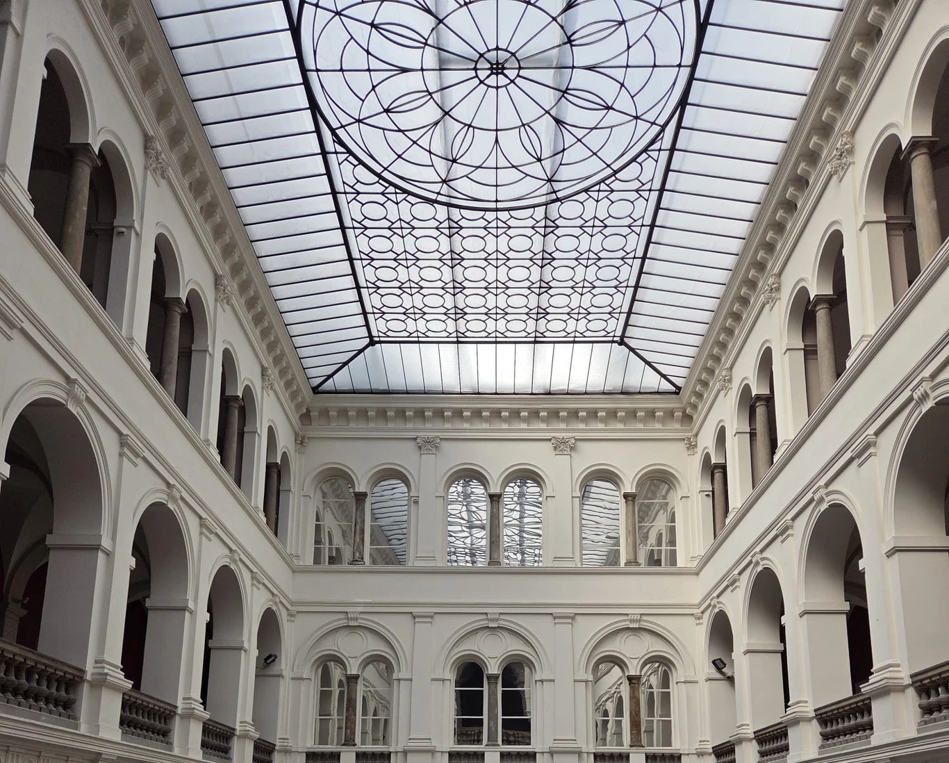 Jasne atrium o klasycznej architekturze z białymi łukami na dwóch poziomach oraz przeszklonym dachem ozdobionym geometrycznym wzorem.