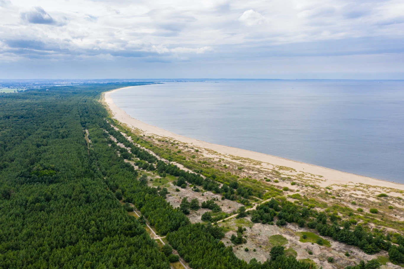 Szeroka, piaszczysta plaża otoczona pasem zielonych lasów, łagodnie przechodząca w spokojną taflę morza pod lekko zachmurzonym niebem.
