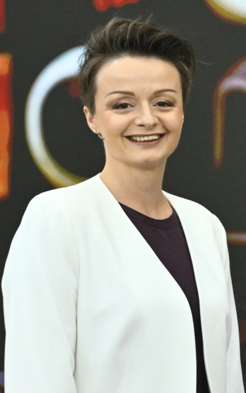 Kobieta z krótkimi, ciemnymi włosami, w białej marynarce i czarnej bluzce, uśmiechnięta, stoi na tle rozmytego, kolorowego napisu.