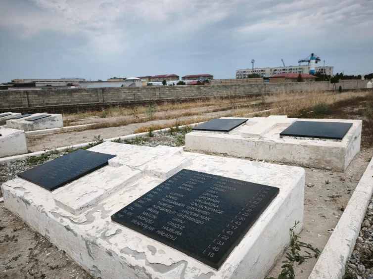 Rzędy białych grobów z czarnymi tablicami nagrobnymi umieszczonymi poziomo, otoczone suchą roślinnością, w tle widoczny mur i zabudowania przemysłowe.