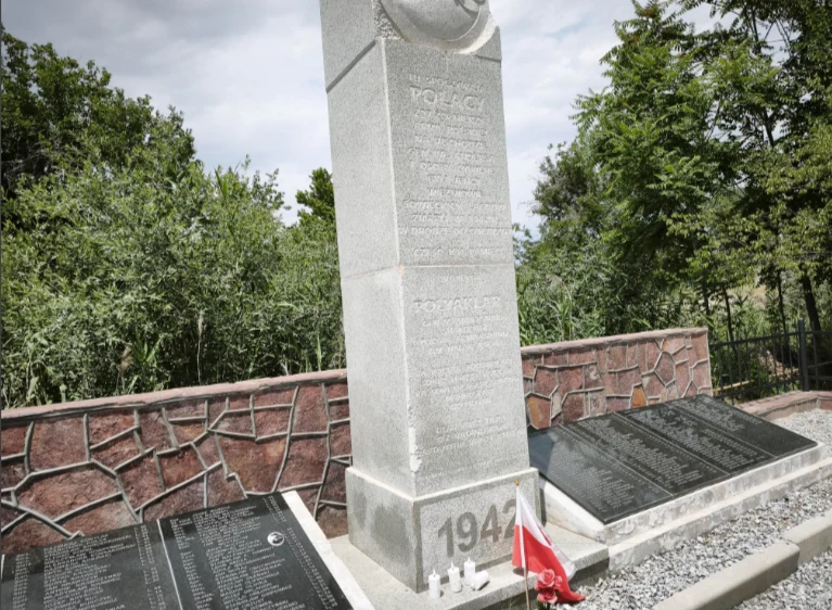 Kamienny pomnik z wyrytą inskrypcją upamiętniający ofiary z 1942 roku, otoczony metalowymi tablicami z nazwiskami oraz małą flagą Polski ustawioną przy podstawie monumentu.