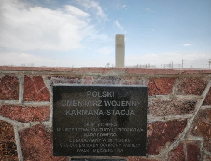 Tablica informacyjna zamocowana na czerwonym murze, upamiętniająca Polski Cmentarz Wojenny Karmana-Stacja oraz wysoki kamienny obelisk widoczny na tle jasnego, częściowo zachmurzonego nieba.