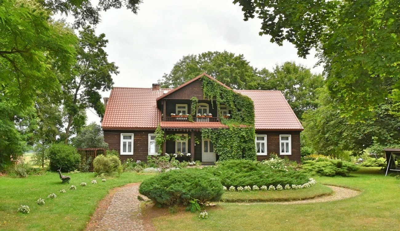 Drewniany dom z czerwoną dachówką, porośnięty pnączami, otoczony zadbaną zielenią oraz ozdobnym ogrodem z krętą alejką z kostki brukowej.